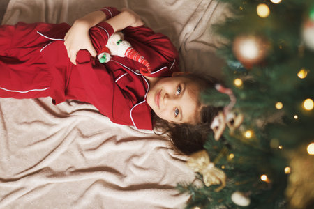 Happy girl of 7 years old in red pyjamas lies on blanket next to Christmas tree with side of garlands and hugging crocheted handmade toy Santa Clausの写真素材