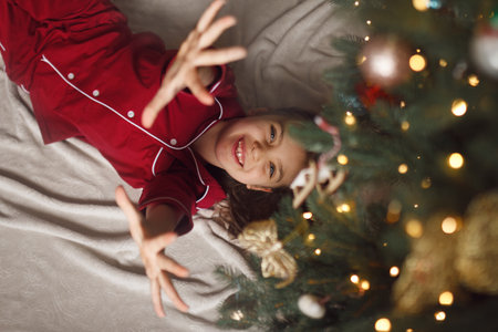 Happy girl of 7 years old in red pyjamas and up in the air lies on blanket next to Christmas tree with side of garlandsの写真素材