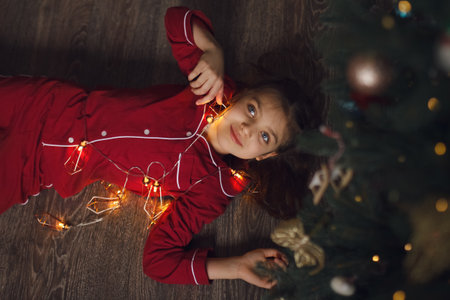 Happy real girl of 7 years old in red pajamas lies with garland of lights under Christmas tree