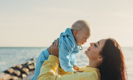 Happy young mother lifted up smiling baby son in blue hoodie on beach with sea and sky in...