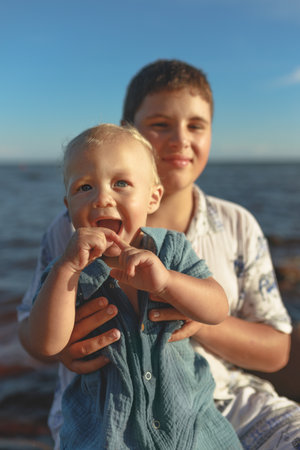 Happy young boy brother holds smiling baby on beach with sea and sky in background