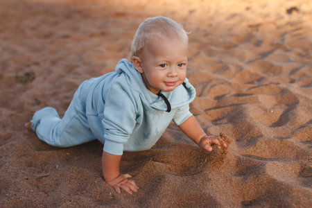 Happy toddler in blue hoodie crawls along beach in sand on sunny dayの写真素材