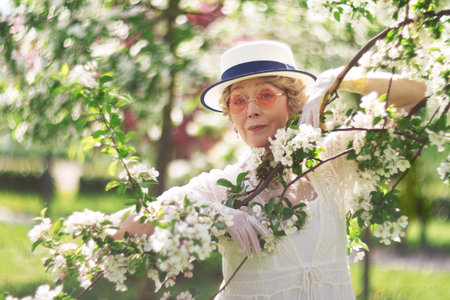 Natural adult mature woman of 60 years old in white canotier hat and glasses closed hugging branch with blossoming apple tree on summer dayの写真素材