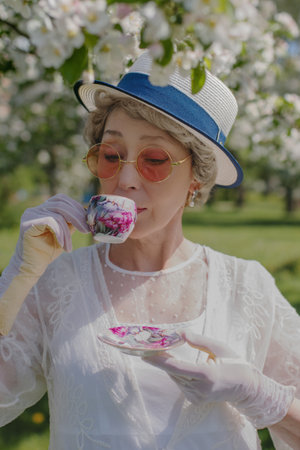 Happy natural adult mature woman of 60 years old in white canotier hat and red glasses, retro dress, gloves drinks tea in blossoming white apple orchard on summer dayの写真素材