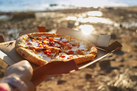 Pizza with tomatoes and mozzarella lies in brown paper box in female hand on beach sun dayの写真素材