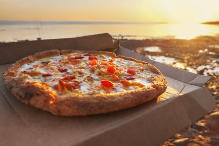 Pizza with tomatoes and mozzarella in brown paper box on sea sunset backgroundの写真素材