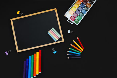 black chalk board and school chalk, felt-tip pens, paints, pen, paper clips, rubber band, on black background top viewの写真素材