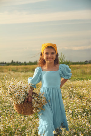 Natural happy girl in blue linen dress and yellow knitted kerchief stands and holds bouquet of flowers in daisy fieldの写真素材