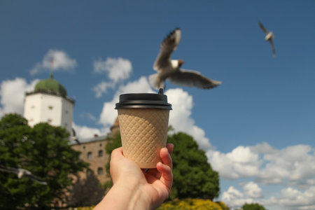 Brown paper cup of drink in womens hand on against backdrop of island Olafs Tower in town Vyborg and...