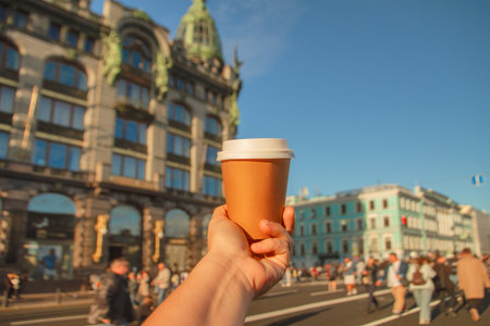 Paper cup of natural coffee in womens hand against backdrop of Singer House on Nevsky Prospekt in center of St. Petersburg, Russiaの写真素材