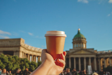 Brown paper cup of drink in womans palm on background of Kazan Cathedral in St. Petersburg and sky on sunny day, copy spaceの写真素材