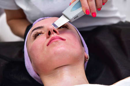 The beautician performs a facial cleansing procedure. Facial care, mask, scrub. Hardware facial cleansing. Skin rejuvenation and beauty.の写真素材