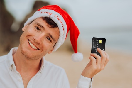 happy smiling and laughing handsome boy men in red hat Santa claus and white shirt stay on beach and hold credit card in oceanの写真素材