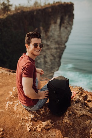 Young man using a backpack on top a ocean cliff, happy smiling emotionsの写真素材