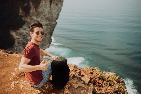 Young man using a backpack on top a ocean cliff, happy smiling emotionsの写真素材