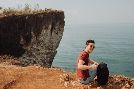 Young man using a backpack on top a ocean cliff, happy smiling emotionsの写真素材