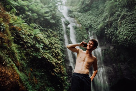 young man muscular and beautiful standing near the waterfall with cellphoneの写真素材