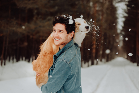 cheerful cute laughing and smiling guy in jeans clothes with dog border collie red on his hands in snowy forest. concept of winter . winter vacationの写真素材