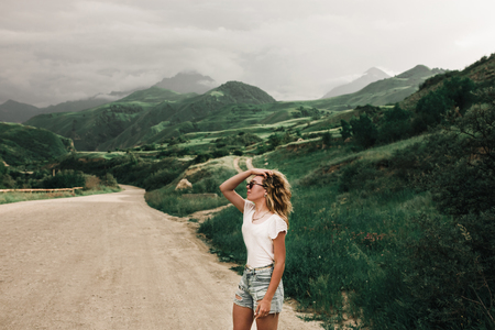 fashionable girl in white clothes standing on the road in the highlands . green grass and mountainsの写真素材
