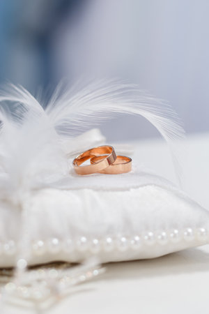 Gold wedding rings on decorative pillow with pearls and white feathers, wedding ceremony blue background, close-upの写真素材