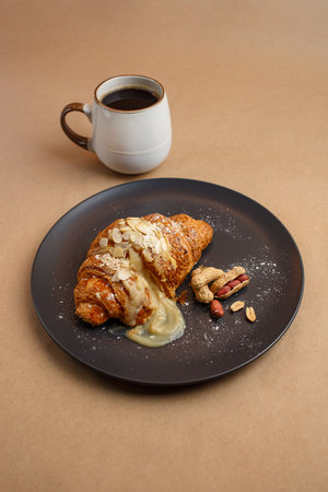 Croissant with sweet filling peanuts and almonds with powdered sugar lies on black plate. Food photo jug coffee on paper brown background, top viewの写真素材