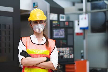 Portrait of female Engineer wear face mask with safety vest and yellow helmet standing in CNC...