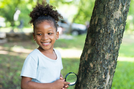 Smiling African American little girl has fun holding magnifying glass to explore and look bugs on the tree between learning beyond the classroom. Education outdoor concept.の写真素材
