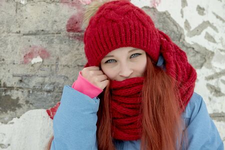 Portrait of young beautiful woman in red hat and red comforter standing against grunge concrete wallの写真素材