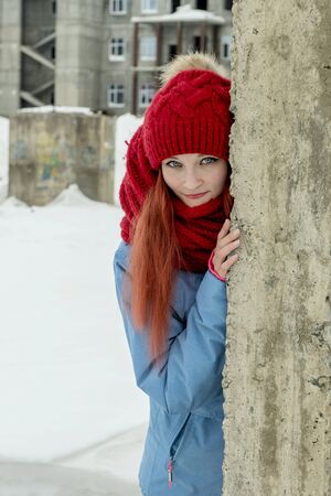 Portrait of young beautiful woman in red hat and red comforter hiding behind grunge concrete wall の写真素材