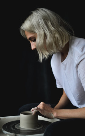 Close up girl ceramist works behind a potters wheel in profile on a dark backgroundの写真素材