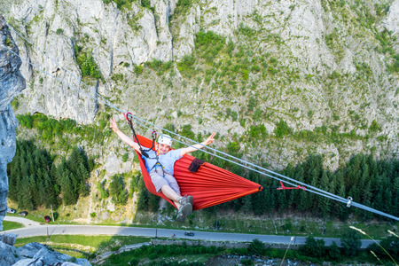 Relaxed tourist male in a hammock high up in a mountain, above the valley, secured with a via...