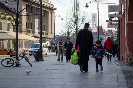 Sibiu, Romania - March 26, 2019: Priest dressed in black walking, holding a child's hand and carrying a bag with groceries, while the kid holds the receipt. Religios, good deeds, philanthropy concept.のeditorial素材