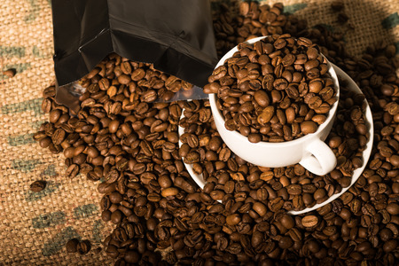 white coffee cup full of beans, surround by more coffee beans and a black bagの写真素材
