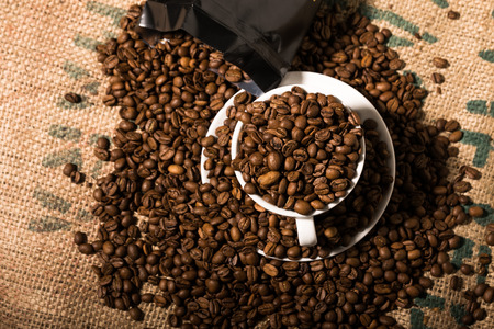 white coffee cup full of beans, surround by more coffee beans and a black bagの写真素材