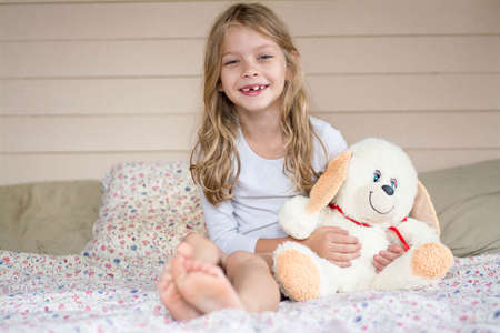 A girl with a plush hare sits on the bed and smiles. Her hare is white. The girl is wearing a white...