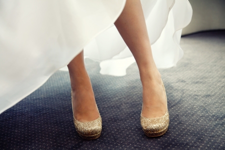 A beautiful bride in a white wedding dress stands outdoors with her pretty gold wedding shoes on her feet の写真素材