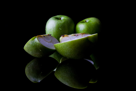 Two whole green apples and one sliced on black background directly from side with reflectionの写真素材