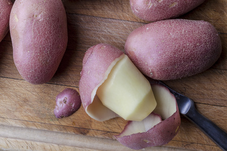 Partly peeled potato with intact ones with peeling knife on wooden cutting board from aboveの写真素材
