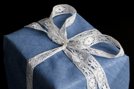 Closeup on white lace ribbon bow tied on wrapped in blue textured paper gift on black backgroundの写真素材