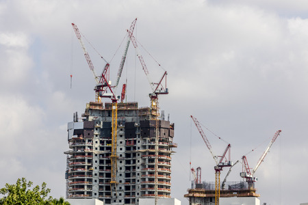 Two buildings one multistory under construction with scaffolds and cranes on top of it green tree cloudy skyの写真素材