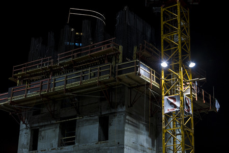 Building under construction stories with yellow crane mast attached on the side lighted by spotlight at nightの写真素材