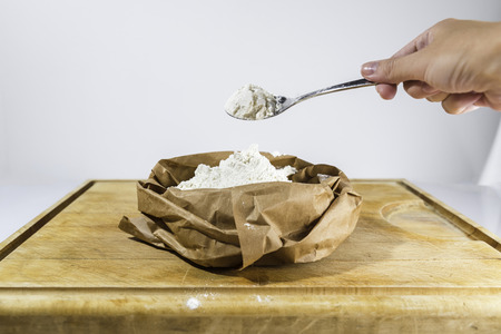 Woman hand holds a spoon of white flour above brown paper flour sack on wooden cutting board on white background from sideの写真素材