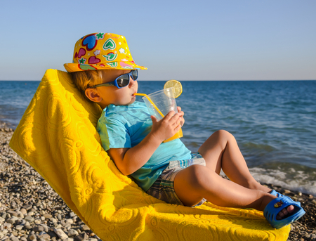 Boy kid in armchair with juice glass on beach against seaの写真素材