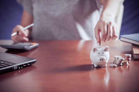 Female hand putting money into piggy bank with using calculator to calculate and money stack, for saving money wealth and financial, business, investment, retirement, saving for the future concept.の写真素材