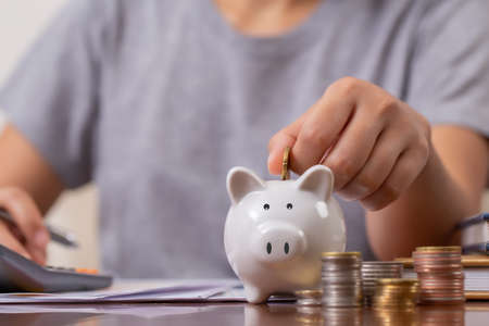 Female hand putting money into piggy bank with using calculator to calculate and money stack, for saving money wealth and financial, business, investment, retirement, saving for the future concept.の写真素材