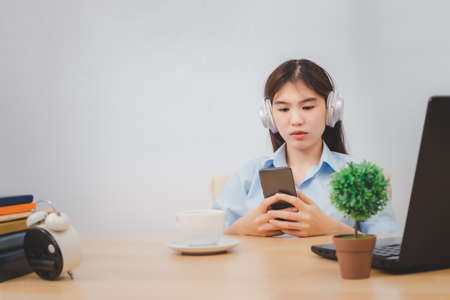 Asian female happy cute beautiful business woman sit indoors in office using phone listening music with earphones.の写真素材