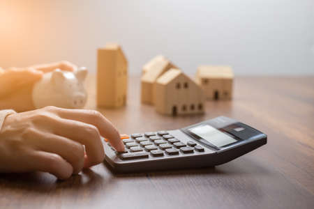 The hand is pressing calculators, piggy bank with wooden house. buy or rent question on note with calculators on desk. Save money and buy house concept.の写真素材