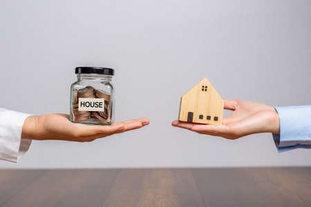 Client hand holding a money jar and house model in home broker hand or salesman person. on wooden...