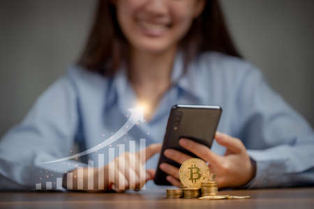 Gold bitcoin and coin pile on wooden table with graph growing and woman use mobile phone as...