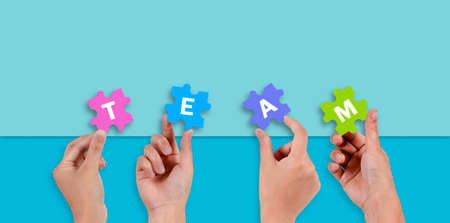 Close up of five hands holding jigsaw puzzle with text TEAM on blue background.